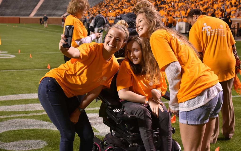 two white females taking a selfie with white female in wheelchair on the football field