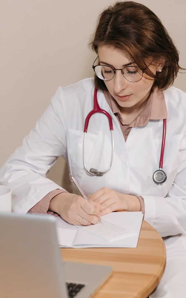 woman in glasses writing on paper at desk wearing white lab coat and red stethoscope around neck