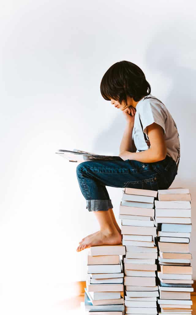 young dark haired student reading while sitting on stacks of books
