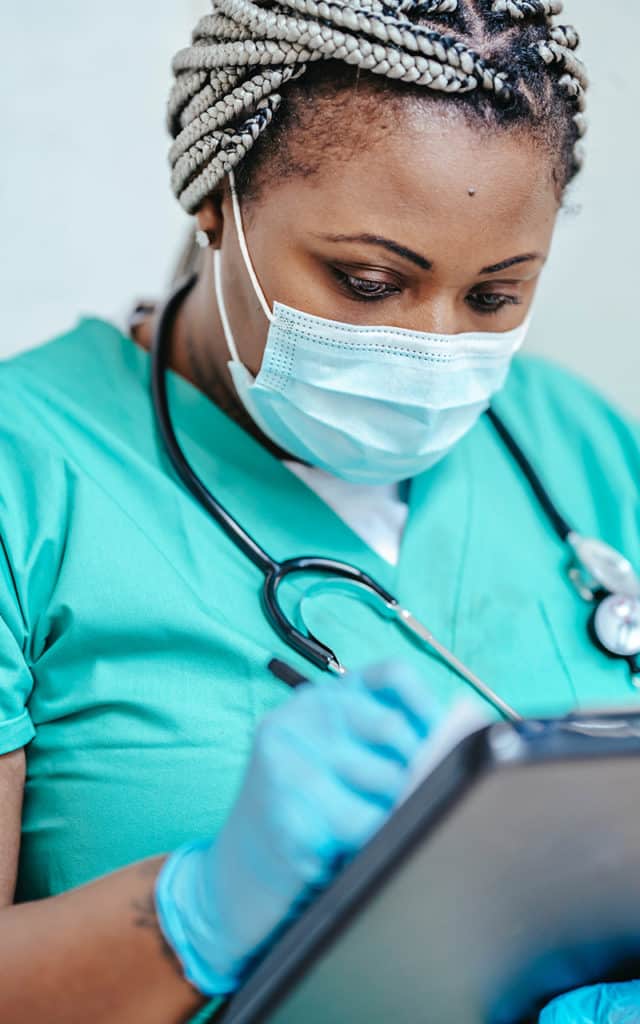 black female nurse wearing green scrubs with black stethoscope writing in patient chart