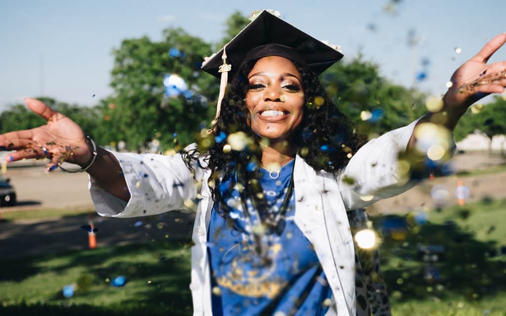 smiling black female nursing graduate with white lab coat and graduation cap throwing confetti 