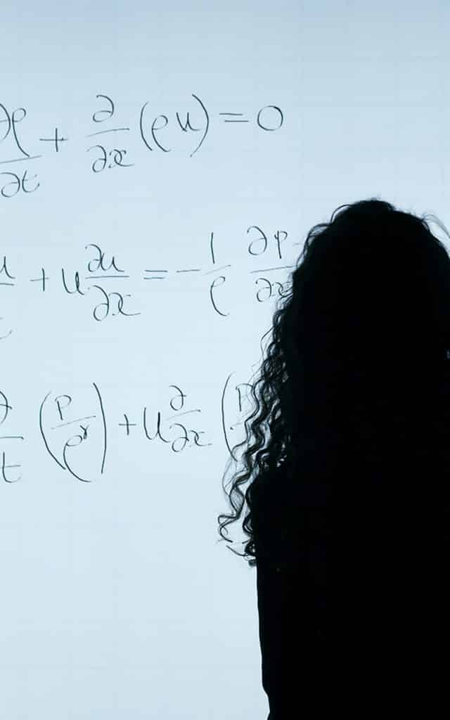 person with long dark hair standing in front of a whiteboard showing mathematical equations