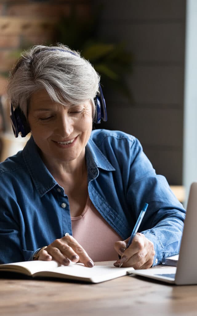older white woman with headphones takes notes during online class